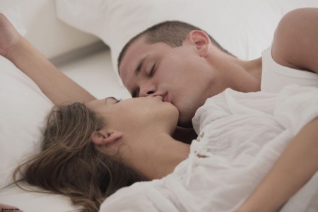 Красивая спящая девушка раскинула голые ножки под одеялом и после поцелуя в спинку порется на боку